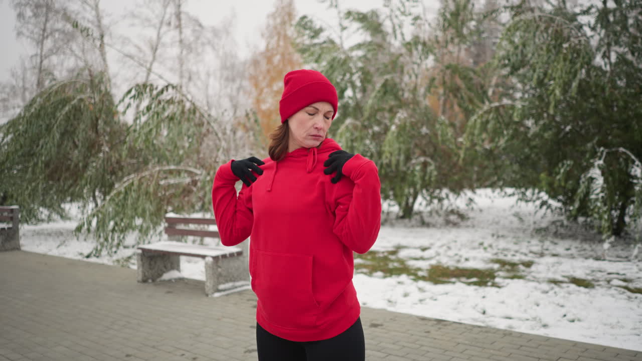 Coach in red beanie stands outdoors with hands on shoulders, performing neck stretches by turning her head left and right in serene winter setting with snow-covered ground, benches, and trees