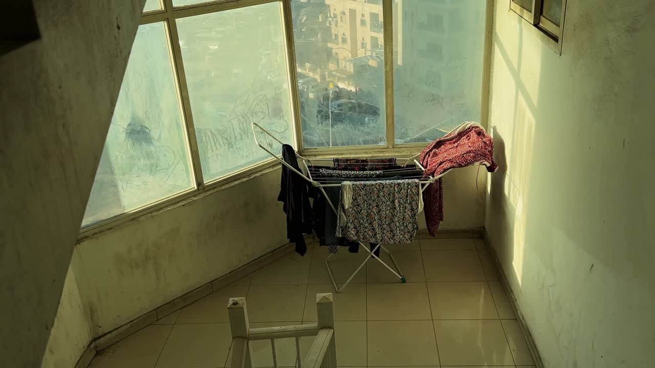 Clothes hang on a folding rack in a sunlit building stairwell landing, warm morning light through large windows, quiet indoor corridor with tiled floor