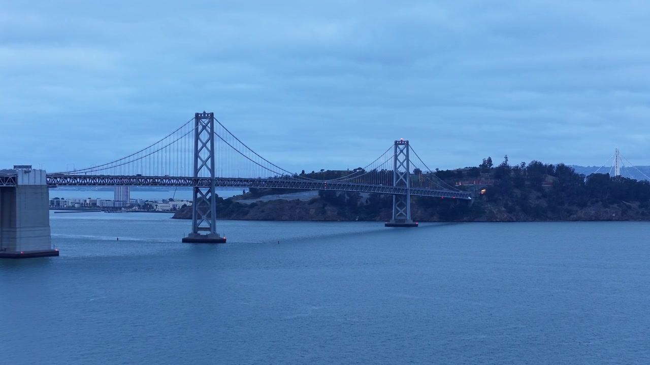 Ampia vista che cattura l'iconico ponte San Francisco–Oakland Bay Bridge che attraversa acque blu al tramonto, con cieli nuvolosi e uno sfondo di isola panoramica, perfetto per temi di paesaggi urbani o di viaggio. Inquadratura ravvicinata con drone.