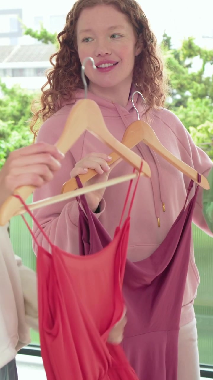 Video verticale: Donna incappucciata che tiene un vestito rosso e ne controlla la vestibilità sul balcone mentre un'amica entra, con spazio per il testo.