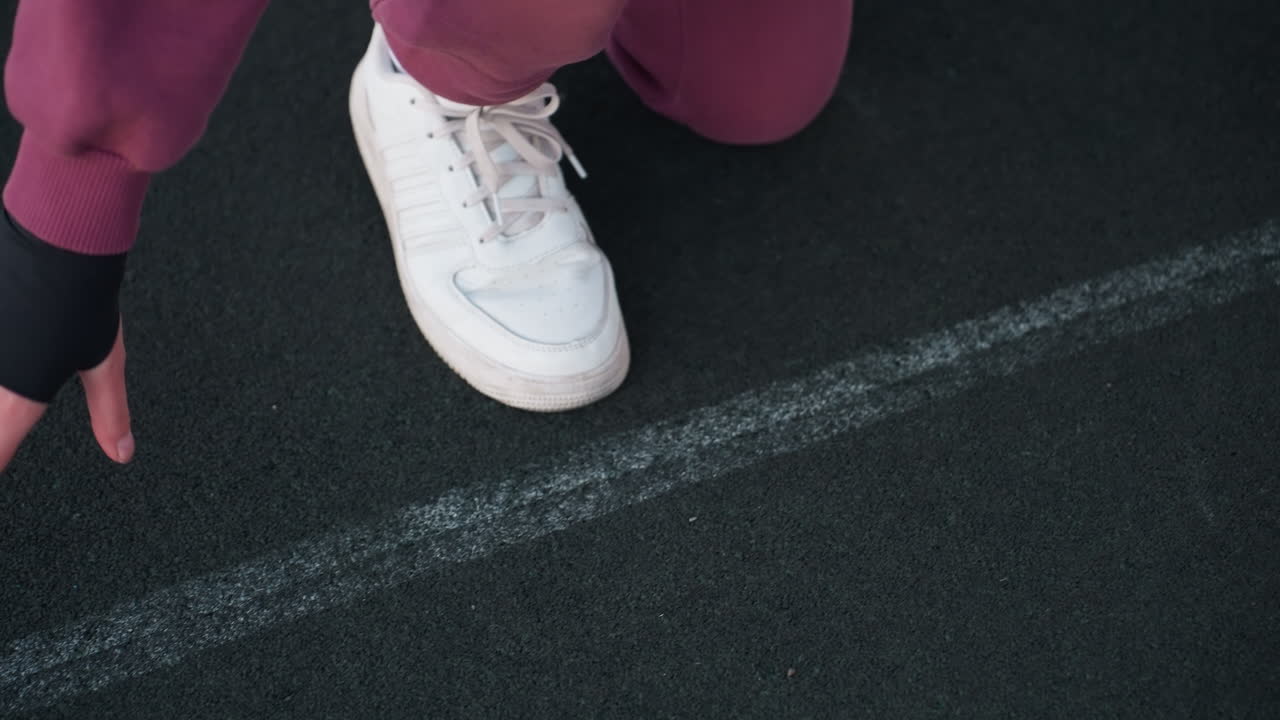 High angle of trainer leg on white court line and hands touching black asphalt surface in urban environment near white barrier topped with chain link fence wearing maroon tracksuit and white sneakers