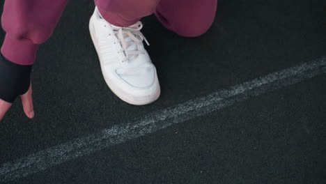 high angle of trainer leg on white court line and hands touching black asphalt surface in urban environment near white barrier topped with chain link fence wearing maroon tracksuit and white sneakers