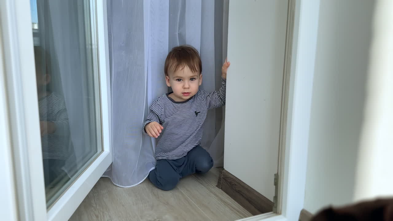 Adorable Caucasian baby boy opens the balcony door and smiles. Black cat at the foreground.