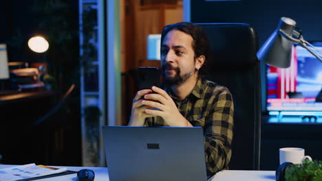 man relaxing at home sitting at office desk, texting friends on smartphone