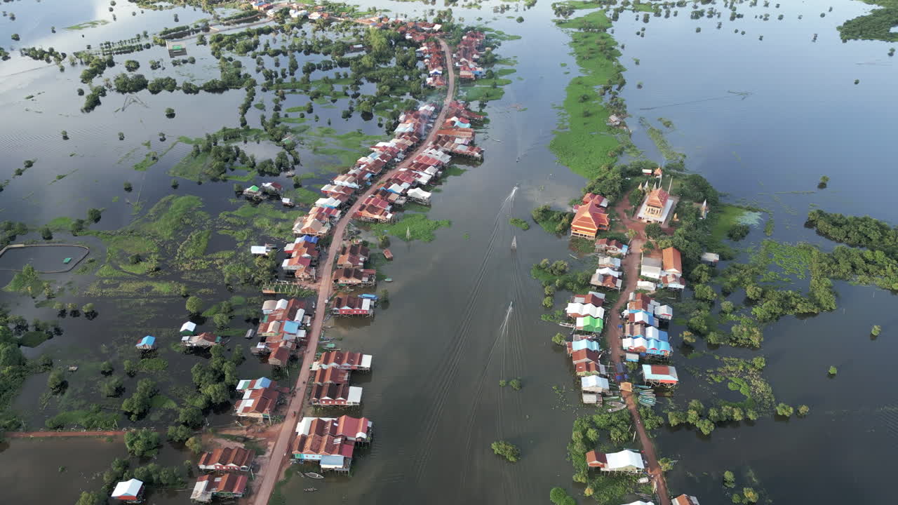 Aerial video of a flooded village in Tonlé Sap, Cambodia. A road divides stilt houses while a temple with bright rooftops stands above the vast water landscape