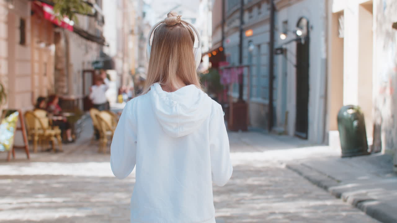 행복한 백인 여성 관광객이 헤드폰을 통해 음악을 듣고 도시 거리를 고 있는 뒷면