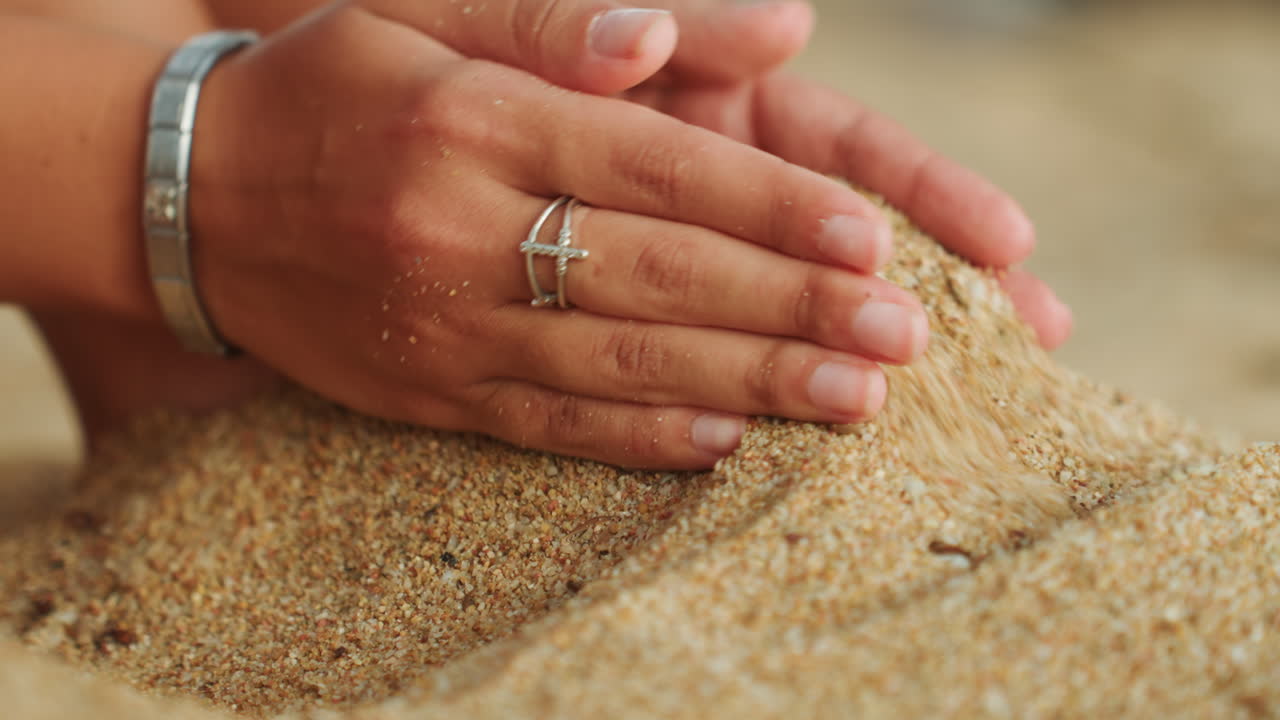 Piedi e mani in primo piano sulla sabbia, texture granulare illuminata dal sole, dita che modellano un piccolo cumulo, anelli e braccialetto visibili, movimento lento e meditativo, atmosfera di vacanza calda, profondità di campo ridotta