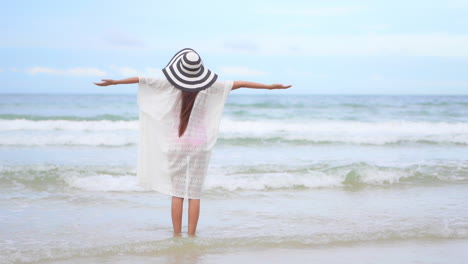 back to the camera a young fit woman in a flopping sun hat and white cover-up welcomes the surf onto the beach