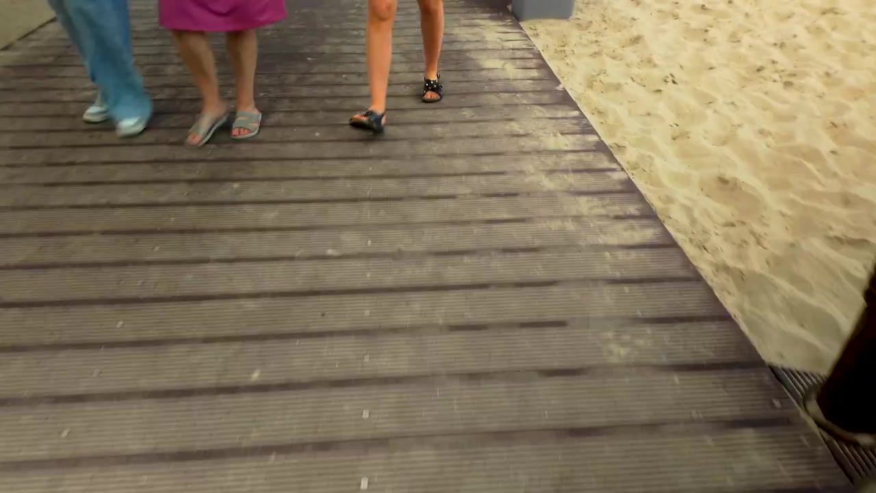 Inquadratura dal basso di turisti che camminano su un sentiero di legno lungo una spiaggia sabbiosa. Con cestini per la raccolta differenziata e infrastrutture costiere