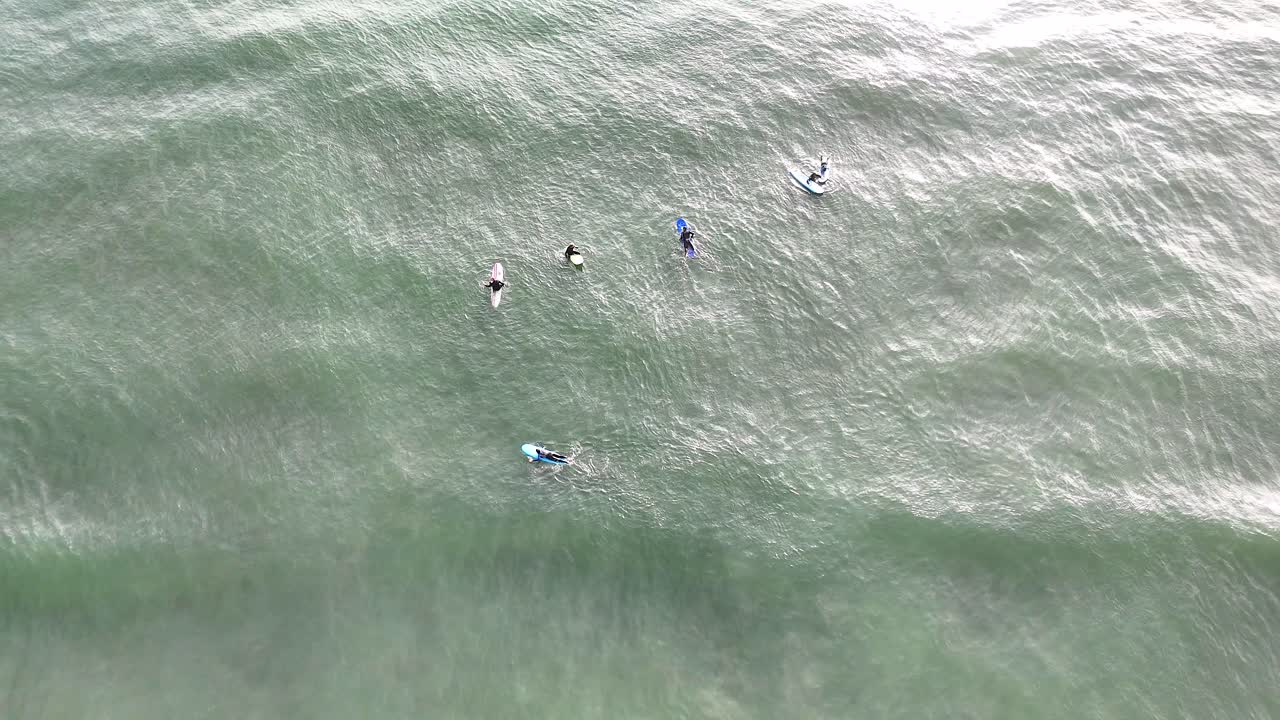 Overhead drone footage shows a group of surfers on boards in calm, sunlit waters off West Sands Beach, St Andrews, Scotland. Minimal movement, tranquil mood