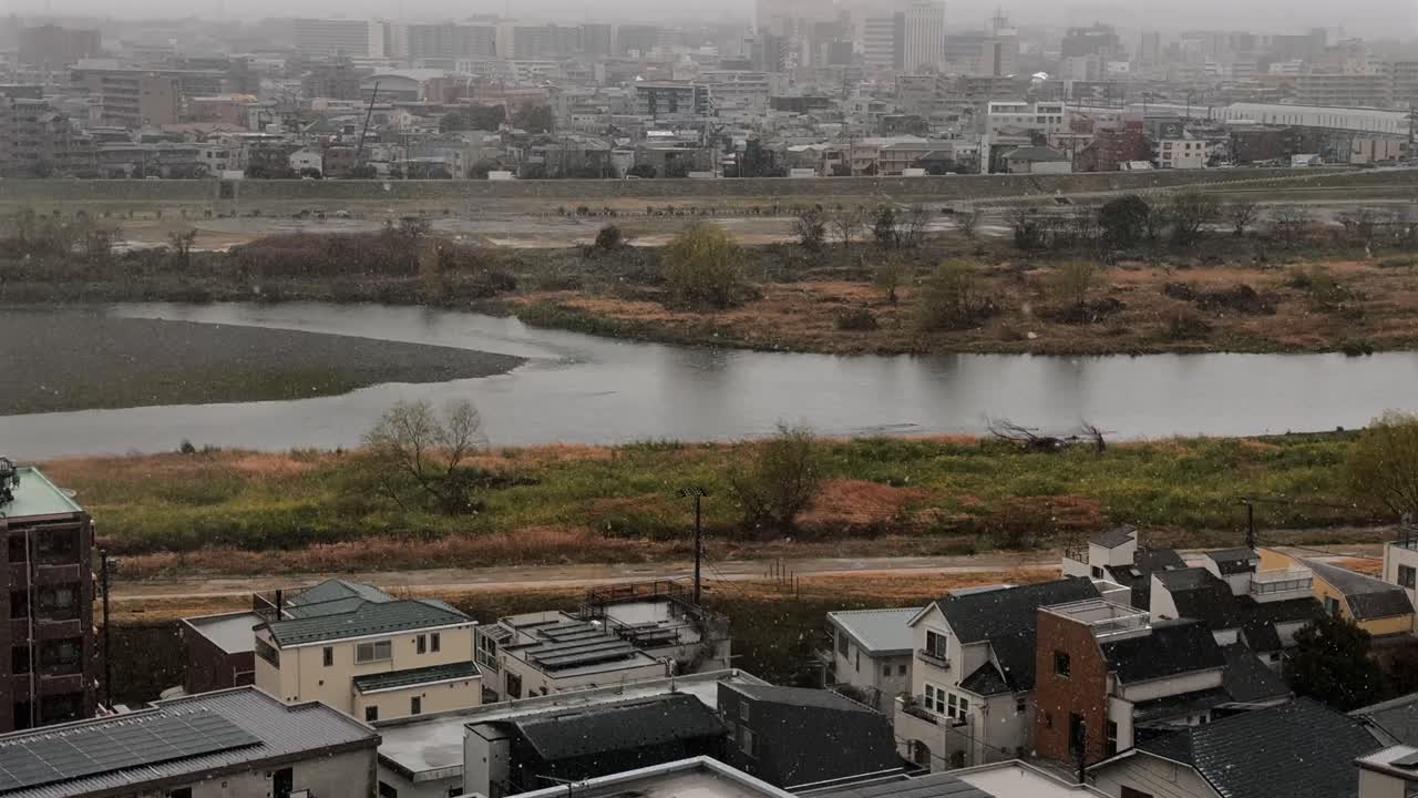 Futakotamagawa on a snowy day, overlooking the Tamagawa River and the neighborhood. Snowflakes drift through the grey, overcast sky, capturing a rare and serene winter scene in Tokyo