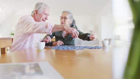 Happy-diverse-senior-couple-sitting-at-table-and-doing-puzzle