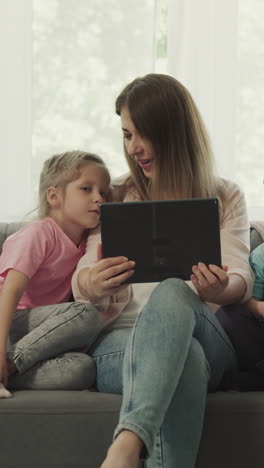 preschooler girl and toddler boy watch mother playing video game on modern tablet. woman enjoys fun activity with children sitting on sofa at home