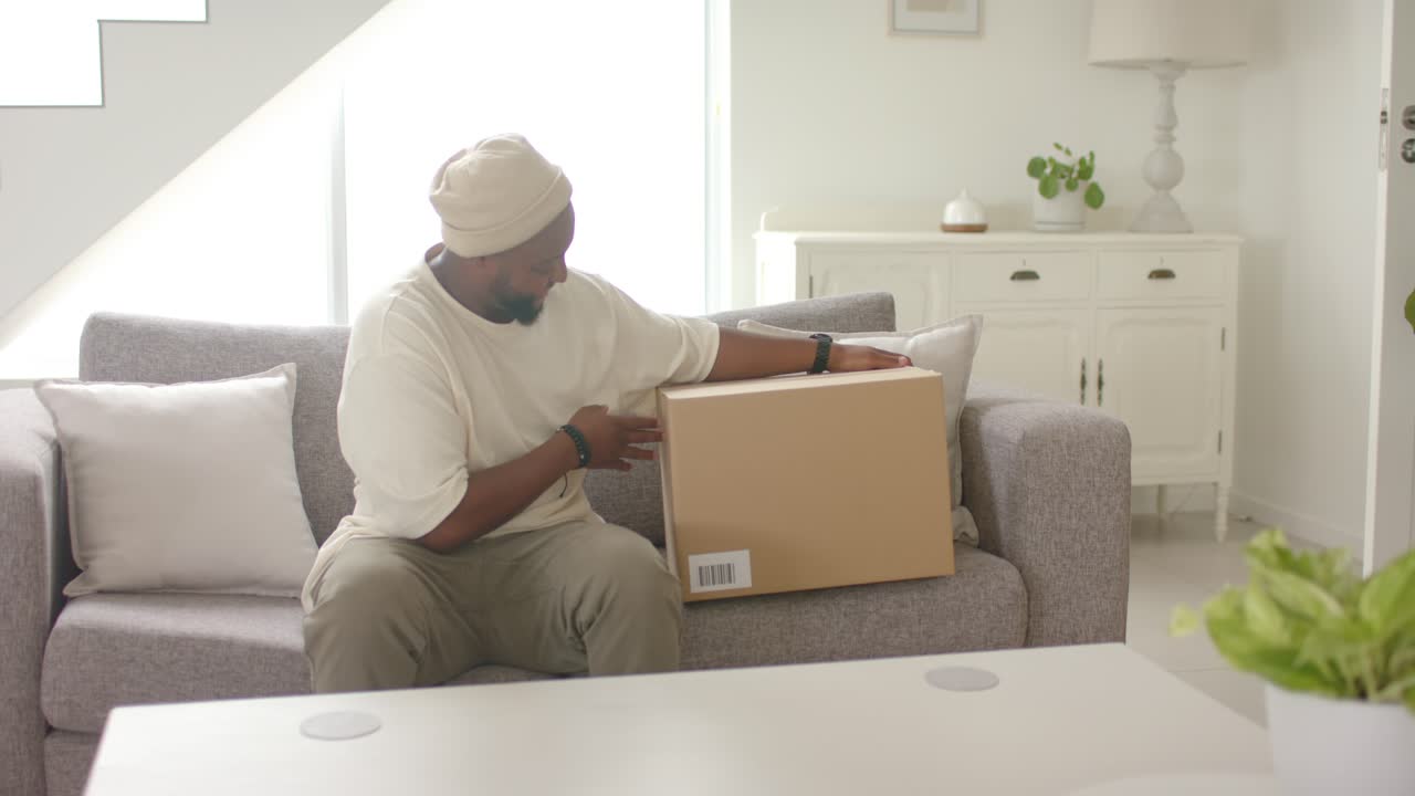 African man placing sealed cardboard box on sofa, steadying, inspecting barcode, preparing to open