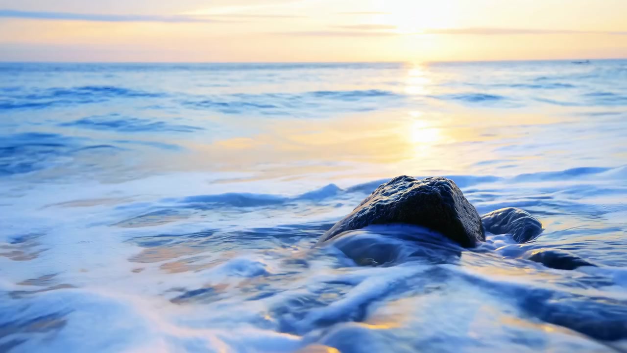 Água do mar espumosa banha rochas costeiras arredondadas, depois recua enquanto ondas suaves se aproximam do mar aberto e a luz dourada se reflete na superfície ondulante.