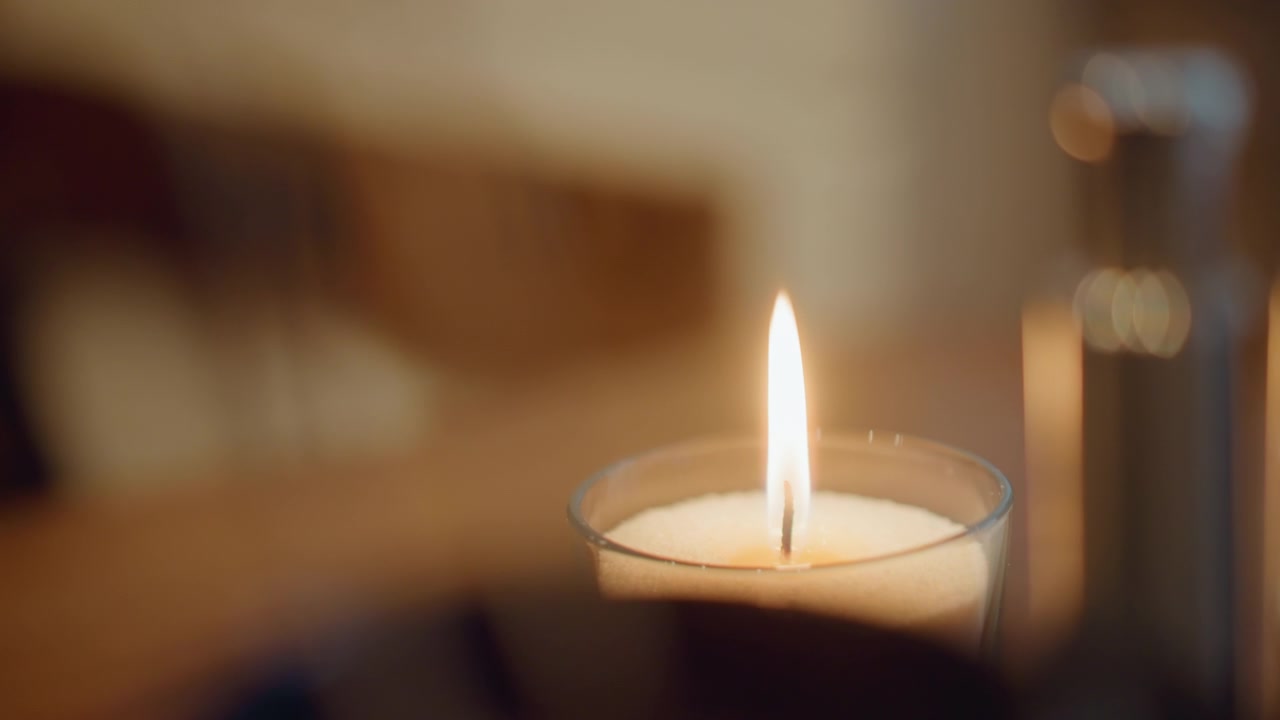 Primo piano di una candela accesa in un bicchiere con fiamma costante e luce calda e soffusa. La cera si scioglie leggermente nel tempo, lo stoppino brucia in modo uniforme in un ambiente interno calmo con dettagli dello sfondo sfocati.