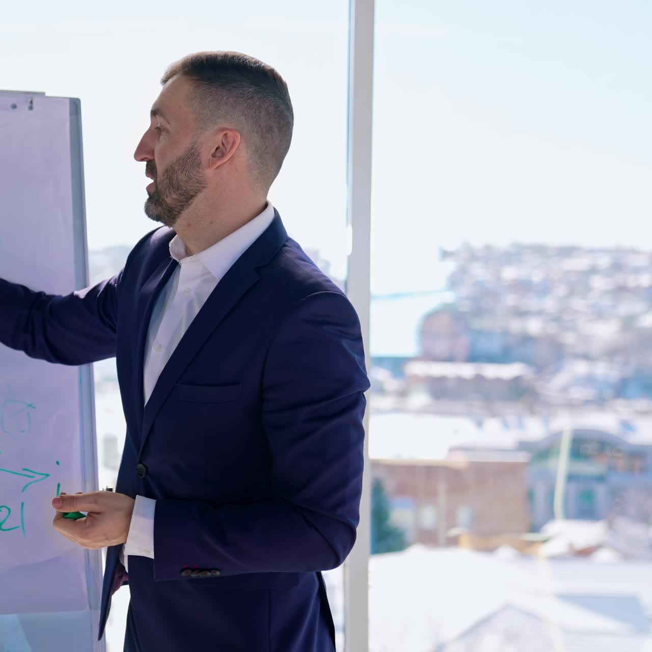 Uomo d'affari che scrive su una lavagna con un pennarello. Imprenditore in abito che parla di problemi finanziari e disegna grafici su una lavagna in ufficio vicino a una grande finestra