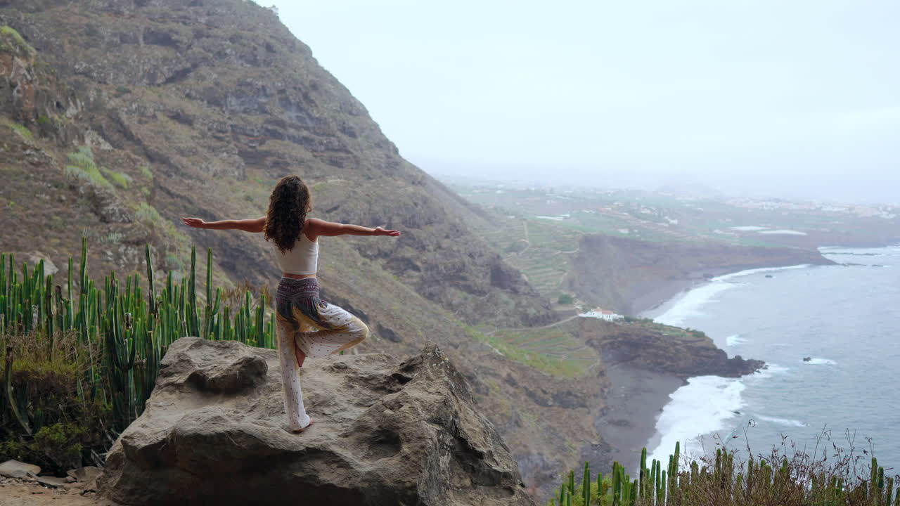 haciendo yoga en un paisaje montañoso de la isla, una mujer joven está de pie en una pierna, los brazos levantados, mirando hacia el océano