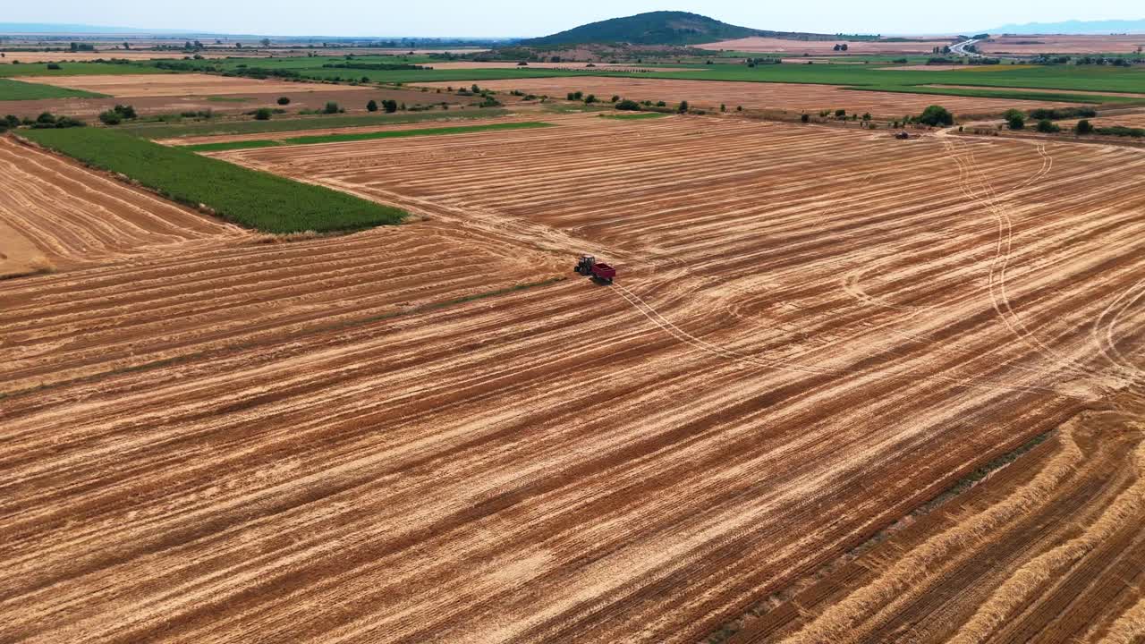 High-altitude aerial shot pushing and curving with a tractor and trailer moving through striped orange farmland, surrounded by endless agricultural fields and distant hazy hills