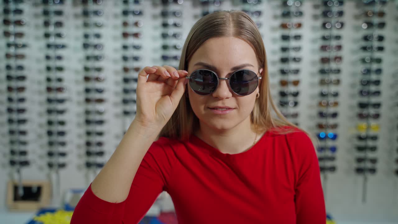 Beautiful woman trying on stylish sunglasses. Long-haired girl in modern black sunglasses posing on camera at optical store.