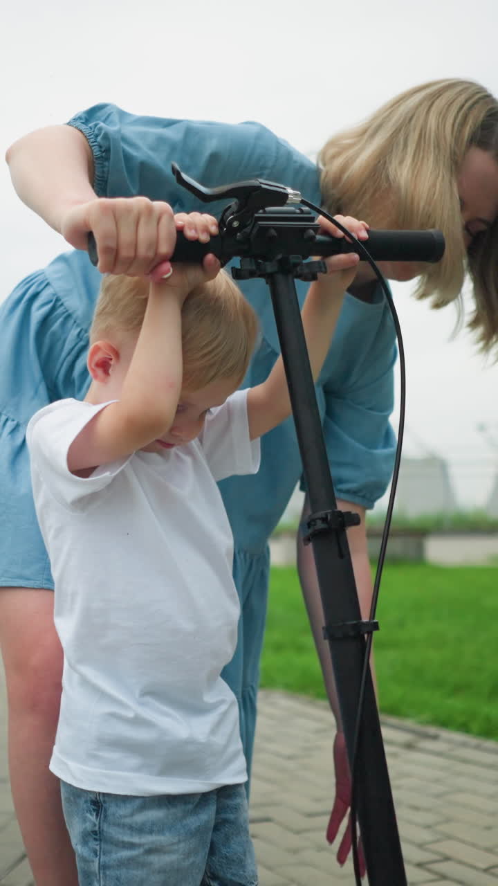 una madre guía cuidadosamente a su hijo sobre cómo pararse correctamente en un scooter, están de pie juntos en un camino entrelazado mientras ella le ayuda a colocar sus manos en el manillar del scooter
