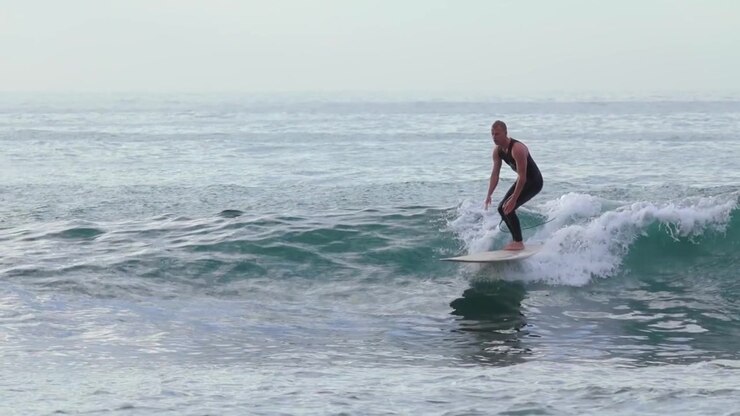 Man wearing a dark wetsuit balancing on a longboard surfboard while catching a small ocean wave near a rocky shoreline. Static eye level view.