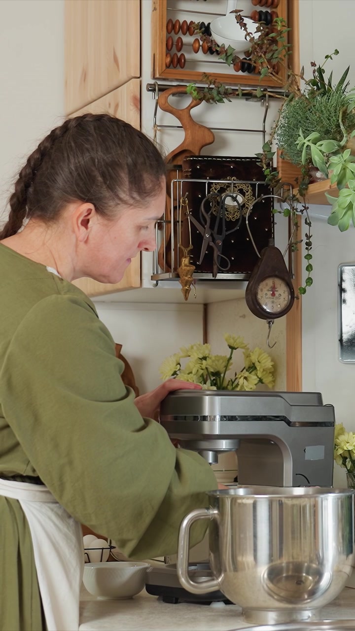 Young woman in green top and white apron operates stand. Woman maintains focus on her task in domestic kitchen. Environment features homey cooking area with wood cabinets