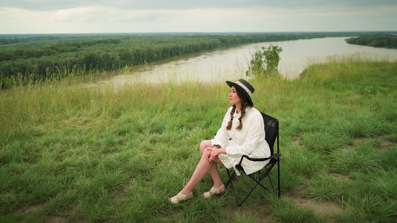 A cute woman wearing a stylish hat and a white dress is seated on a chair in a lush grass field beside a tranquil lake. She poses elegantly with a serene expression under a cloudy sky