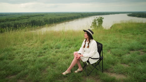a cute woman wearing a stylish hat and a white dress is seated on a chair in a lush grass field beside a tranquil lake. she poses elegantly with a serene expression under a cloudy sky