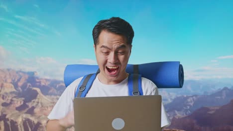 close up of asian male hiker with mountaineering backpack looking at a laptop and screaming goal celebrating the success while traveling at the top of mountain