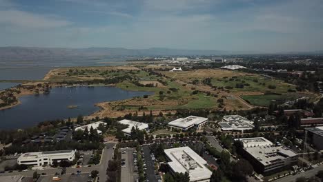 Vogelperspektive-Aus-Der-Vogelperspektive-Auf-Die-Küste-Des-Sees,-Google-Campus,-Amphitheater,-Park,-Berghintergrund,-Schwenk-Nach-Links