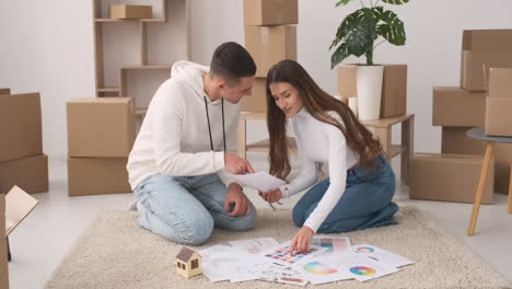 vorderansicht des jungen paares in einem neuen haus, das auf dem teppich sitzt und farben für die dekoration auswählt