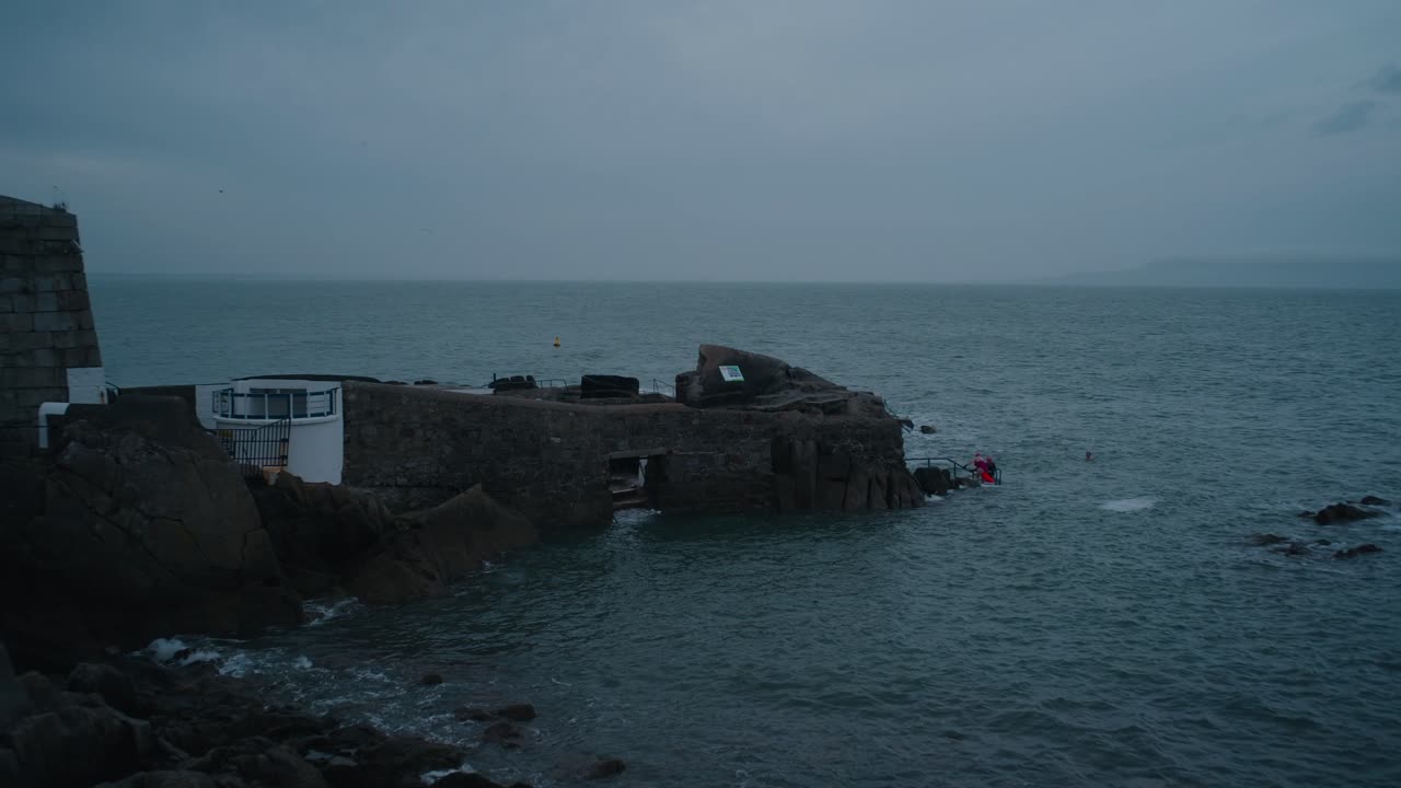 Cold water swimmers enjoying a dip at the Forty Foot in Dublin, a famous Irish sea swimming location. Rugged coastline and overcast sky add to the dramatic scenery.