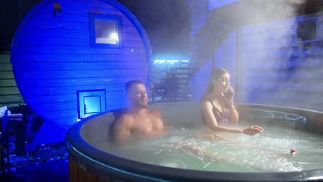 caucasian couple sitting in the hot tub with vapor rising up. long-haired woman speaks on the phone.