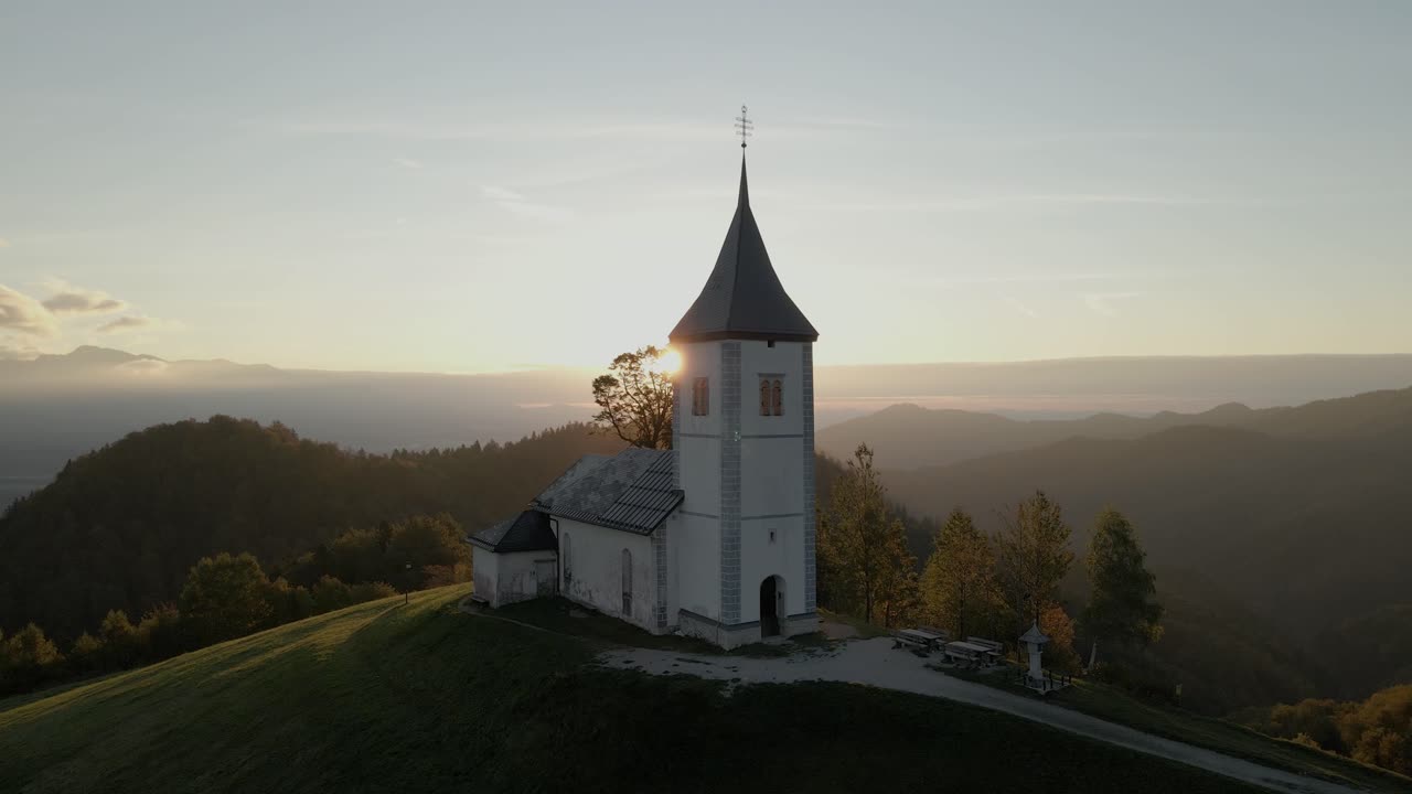 Malerische Luftaufnahme der Kirche St. Primoz auf einem Hügel mit Sonnenaufgang und Berggipfeln, Jamnik, Slowenien