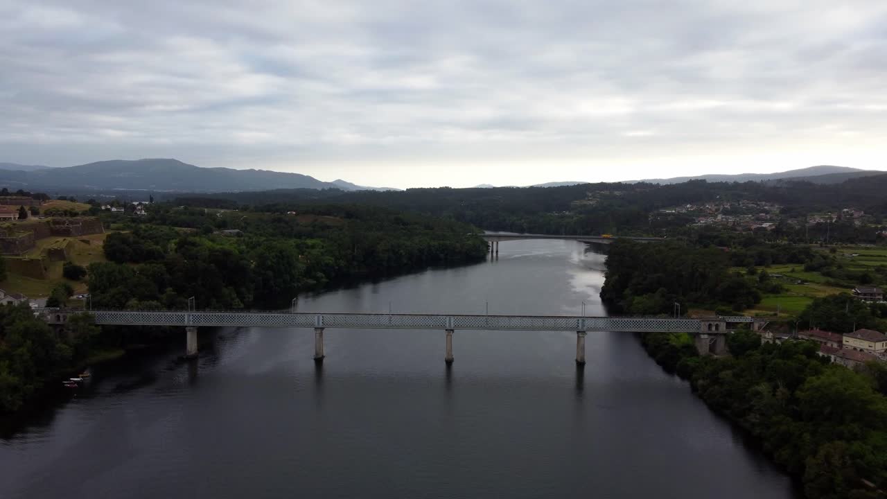 4K drone shot tracking along the Minho River, featuring the main bridge connecting the Spanish and Portuguese sides near Tui, Galicia, with dark water and an overcast sky