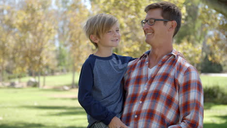 father carrying seven year old son smiles to camera in park