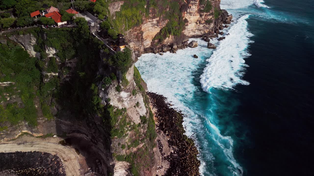 Drone footage of Uluwatu coastline where foamy waves crash below towering cliffs topped with an ancient cliffside temple, revealing the dramatic and vibrant natural beauty of southern Bali