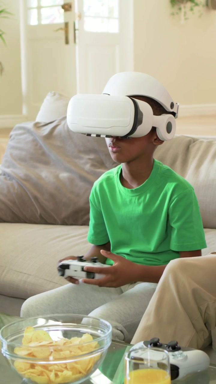 Video verticale: Genitore che porge, posiziona e regola un visore VR sul bambino seduto sul divano del soggiorno per giocare.