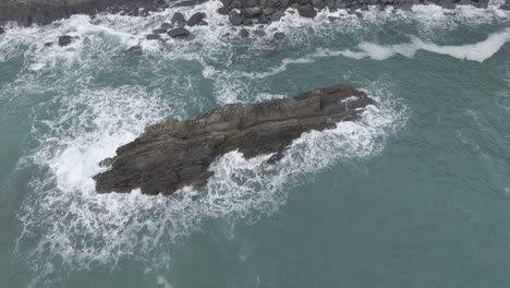 aerial view of a rock jetting our of the water with waves crashing up the side of the rock and birds flying