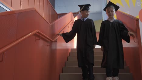 two multiracial kindergarten students in cap and gown talking together while going down the stairs at the preschool graduation ceremony