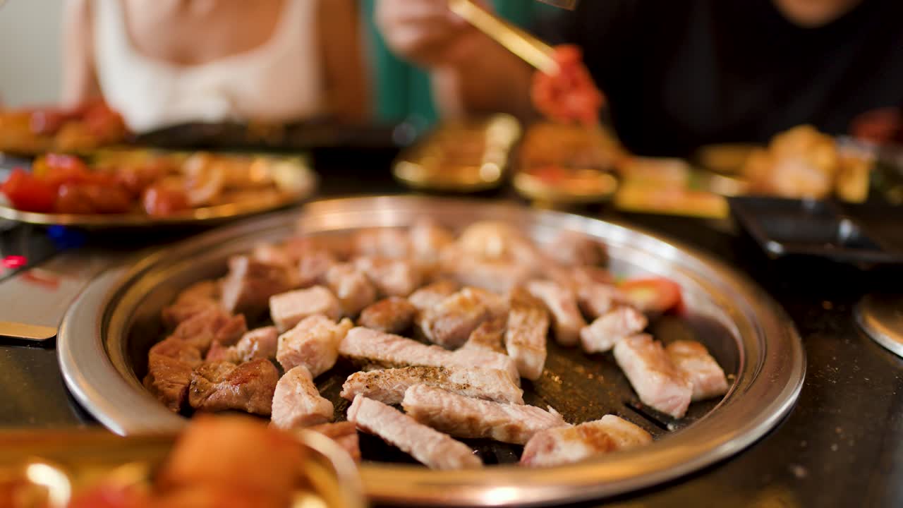 Two people grill pork and chicken on tabletop grill, warm lighting, shallow depth of field