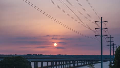 red yellow ball of sun rises through wispy clouds behind utility poles over highway bridge crossing river, time lapse