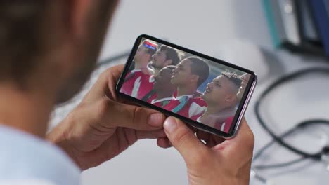 compuesto de un médico masculino viendo un evento deportivo en un teléfono inteligente