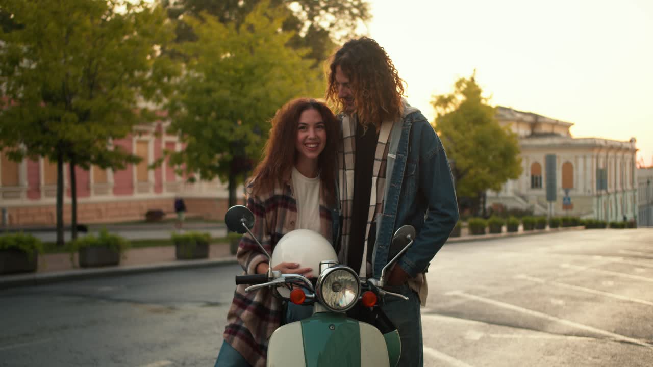 Portrait: happy guy and girl in plaid shirts with curly curly hair posing and looking at camera Near their green moped near a street with trees and ancient architecture on a wide road