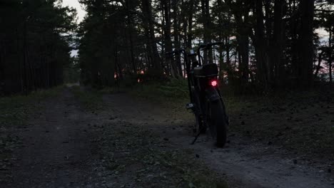 Bicicleta-Eléctrica-Estacionada-En-La-Carretera-Rural-Durante-La-Puesta-De-Sol-Con-Retroiluminación-Roja-Encendida,-árboles-Moviéndose-En-El-Viento