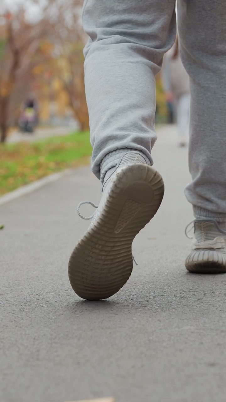 Two legs moving forward wearing grey pants and shoes. Paved surface appears smooth with occasional leaf near foot. Background shows autumn foliage soft focus people in distance