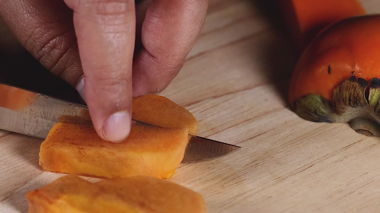 Detailed view of hands slicing an orange fruit into thin pieces on a wooden board.