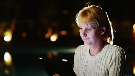 Mujer-Atractiva-Disfruta-De-La-Tableta-Tarde-En-La-Noche-Cerca-De-La-Piscina