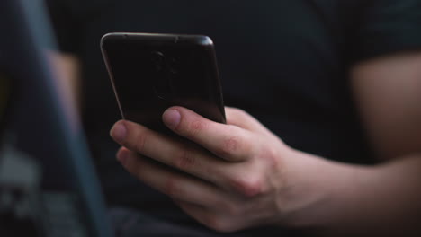 person scrolls news feed on mobile phone sitting at laptop